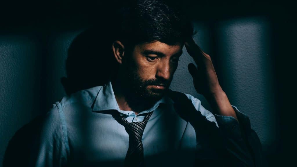 Close-up of a distressed man in a shirt and tie holding his forehead in shadow.