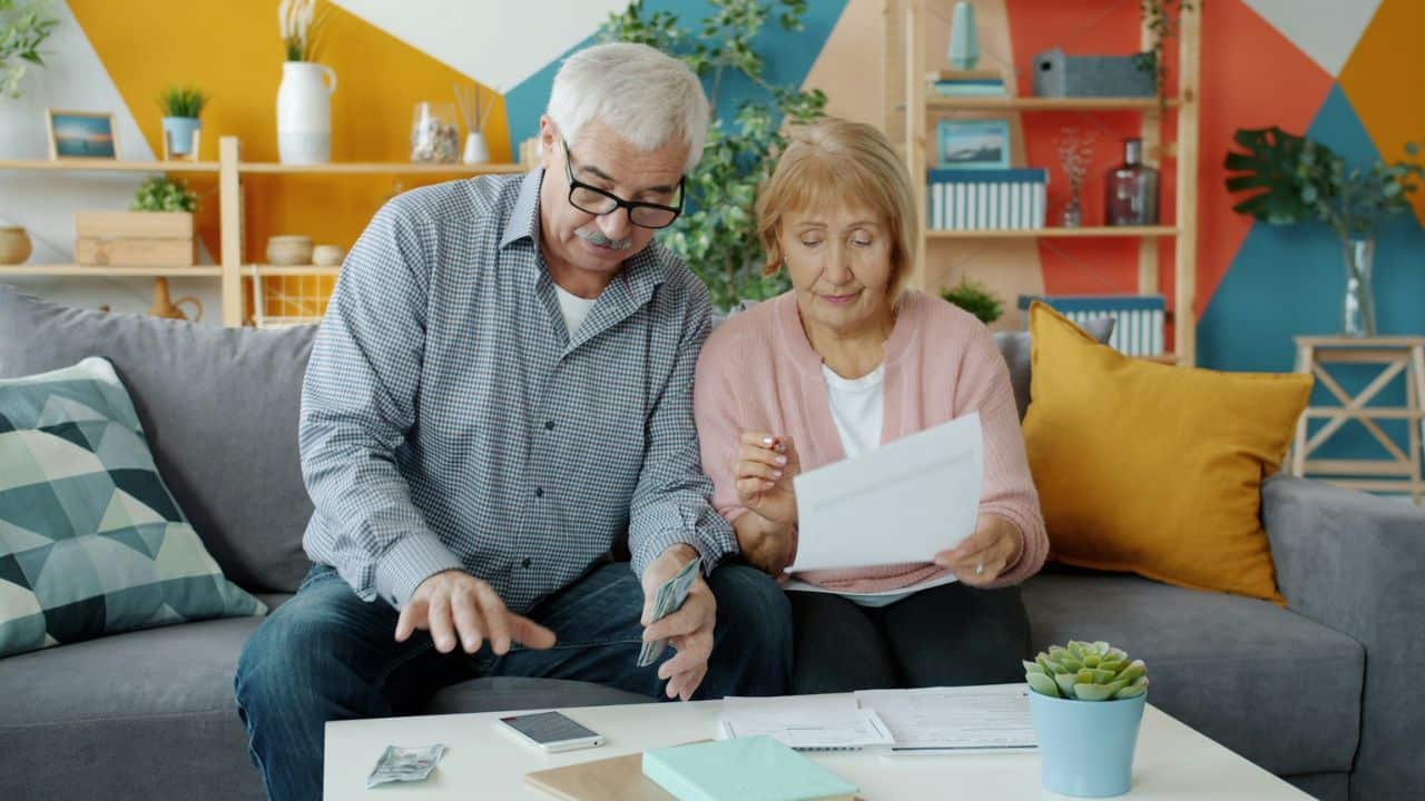 Older man and woman sitting on a sofa, counting money and reviewing papers together.