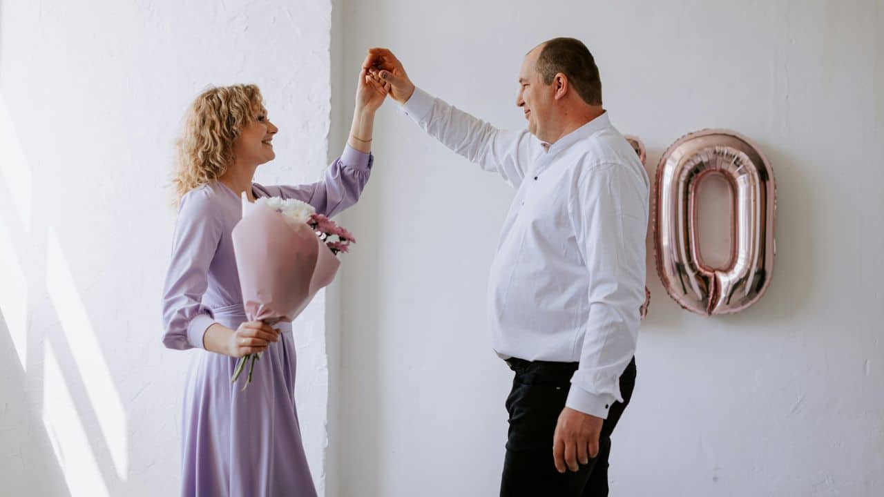 Smiling couple dancing against a white wall with rose gold "00" balloons; woman holds a pink bouquet.