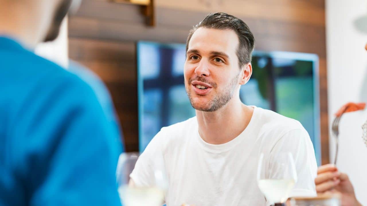 A man in a white shirt smiles while talking to someone at a dining table with wine glasses.