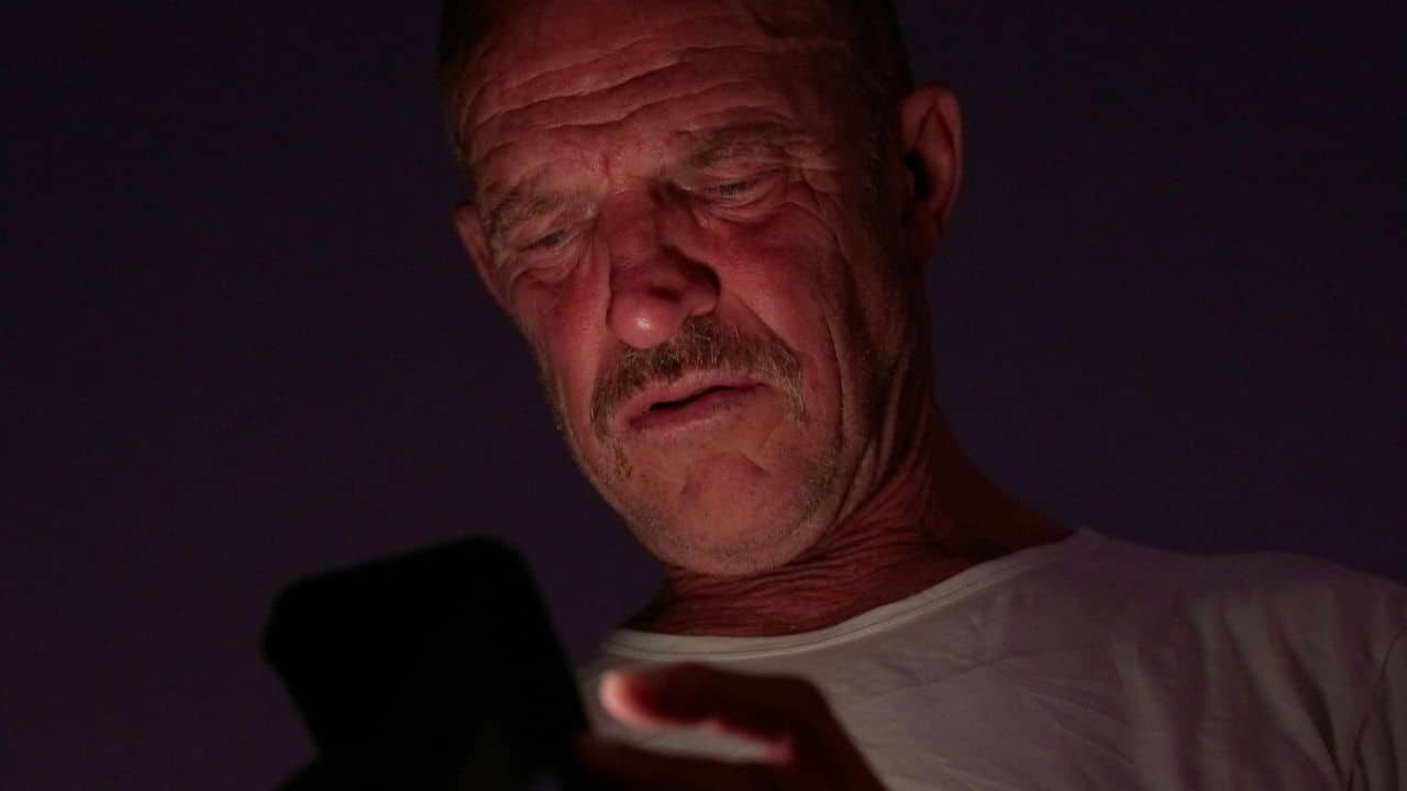 Older man in dark setting looking intently at a glowing smartphone screen.