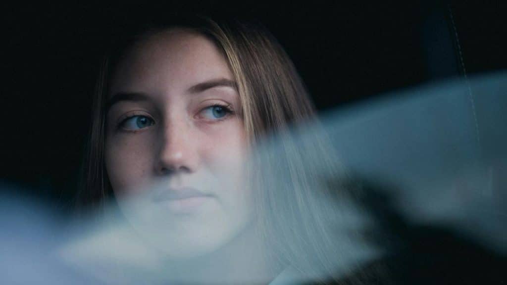 A young woman gazes thoughtfully out of a window with soft light on her face.