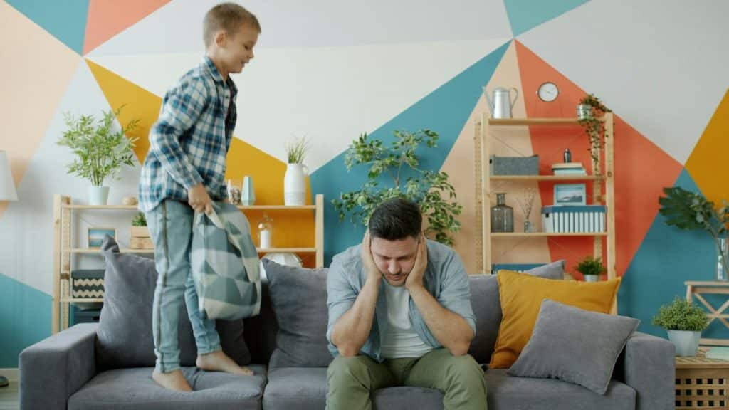 A distressed man sitting on a couch while a boy jumps behind him.