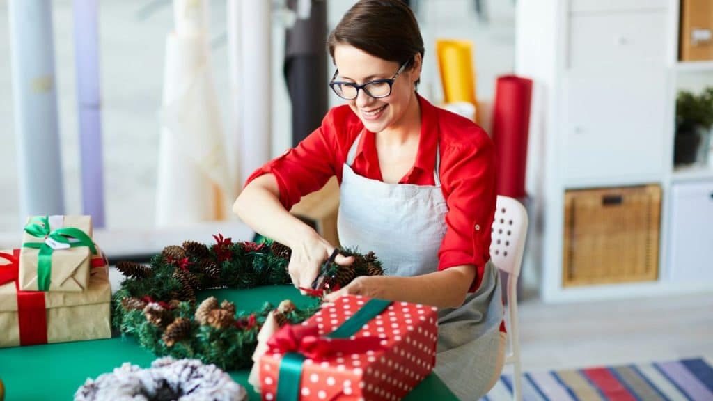 A woman in red and a gray apron smiles while making a Christmas wreath.