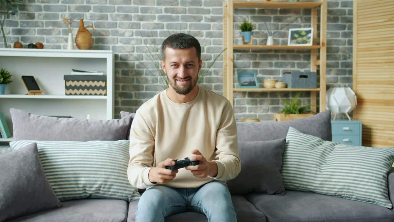 A smiling man sitting on a couch, holding a black video game controller.