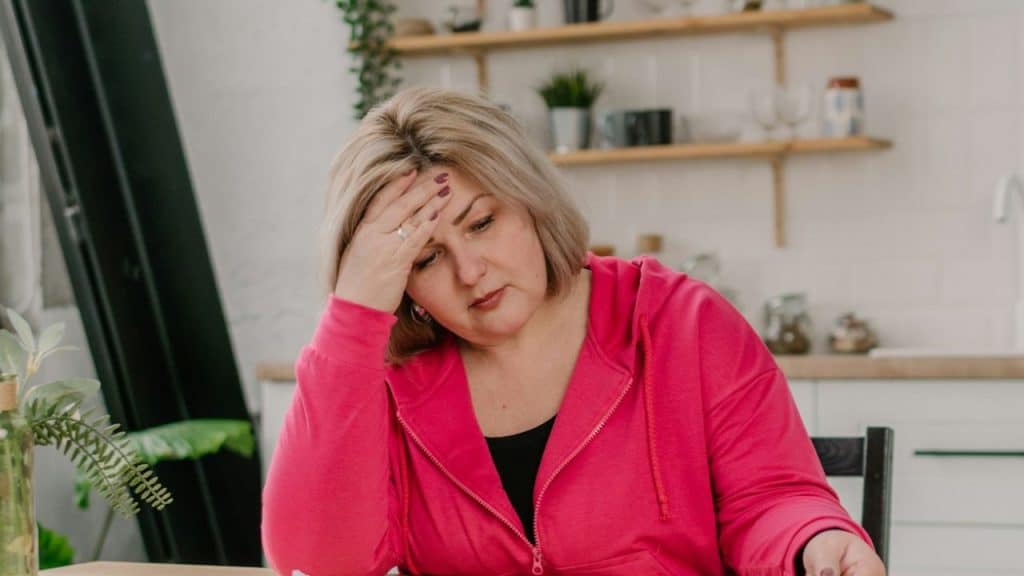 Middle-aged woman in a bright pink hoodie resting her hand on her forehead in a kitchen.