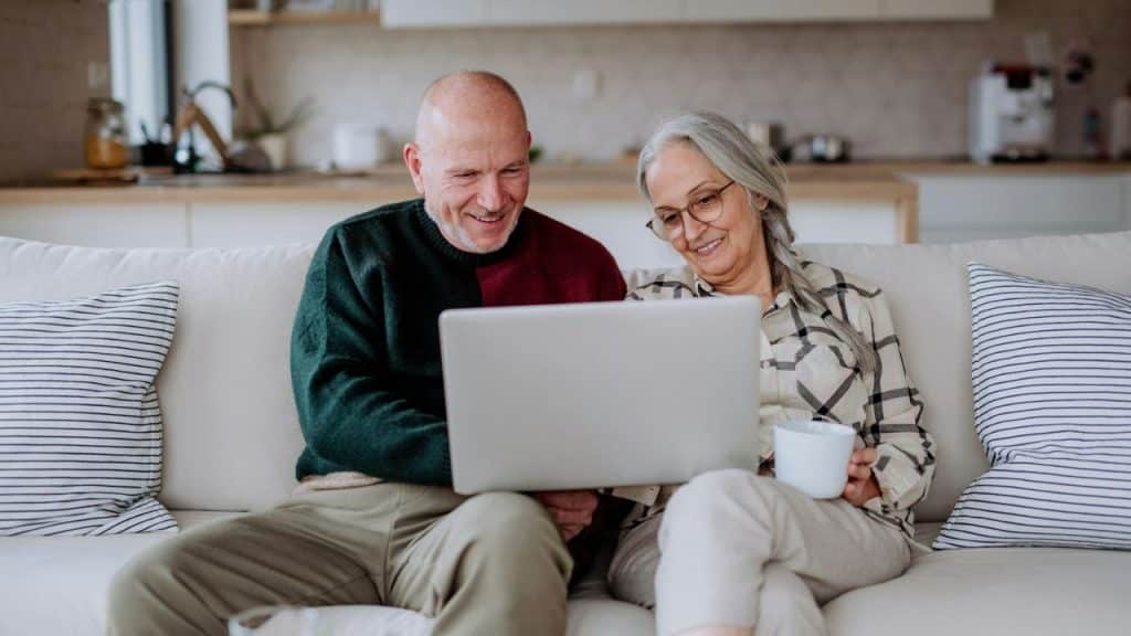 Mature couple sitting on a sofa, smiling while looking at a laptop screen.