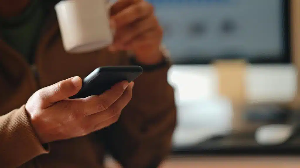 A person holding a smartphone and a white mug while sitting at a desk.