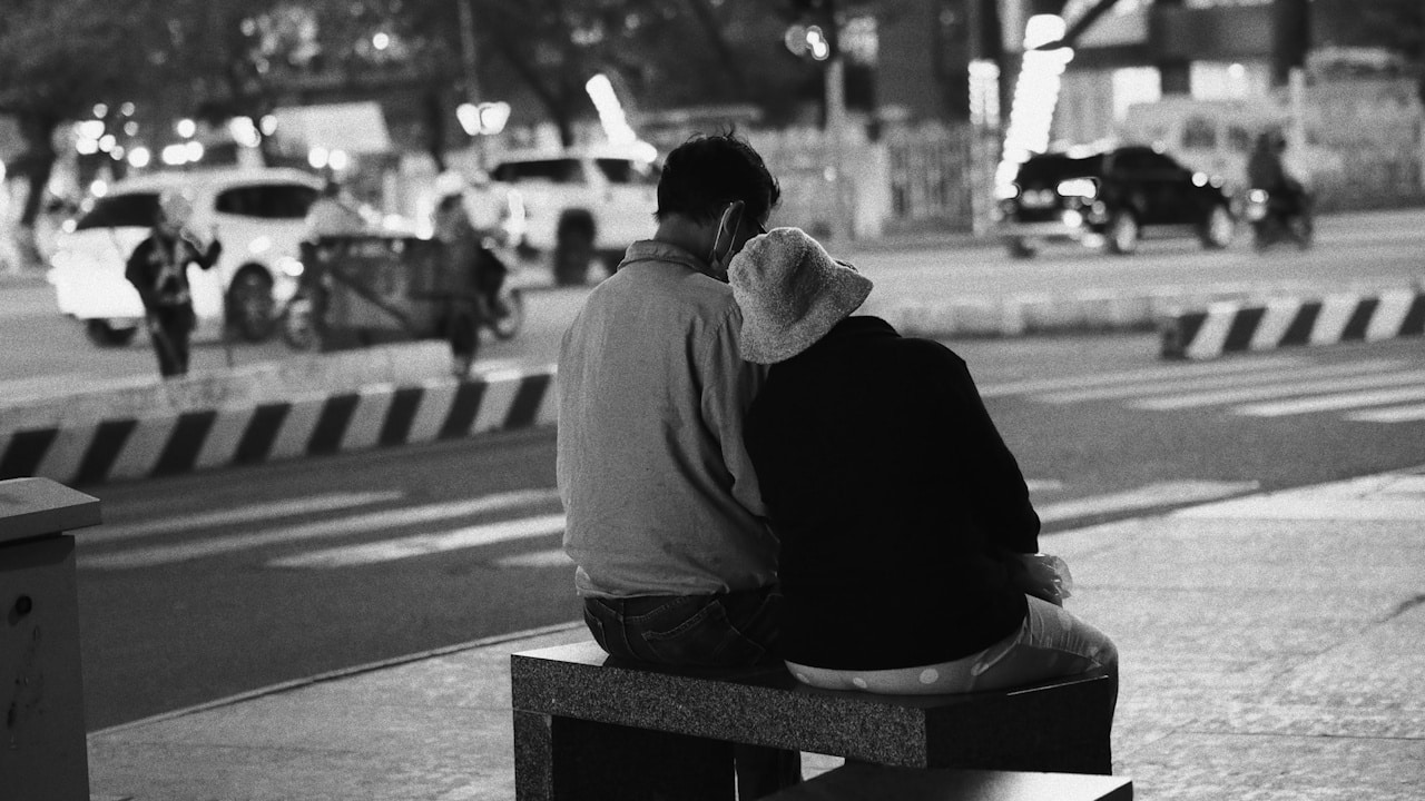 A black and white image of a couple sitting close together on a bench by a street at night, seen from behind.