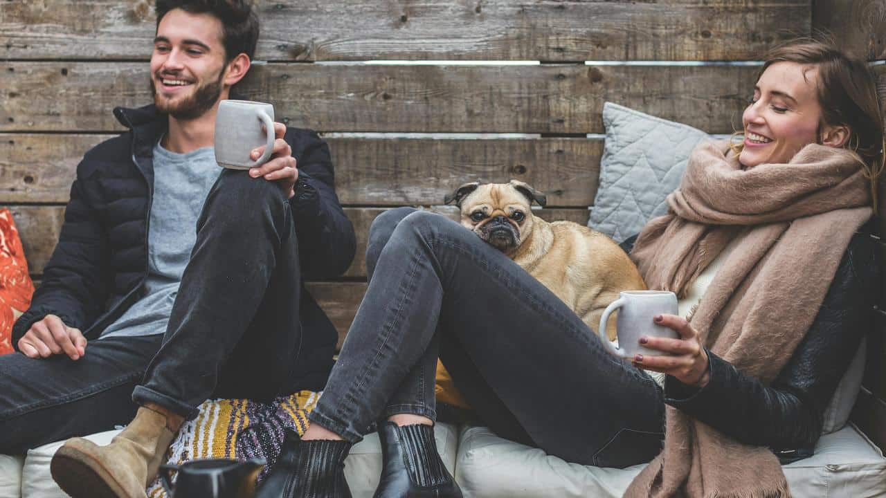 A couple relaxing with hot drinks and a small dog between them.