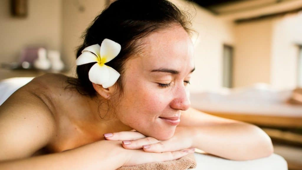 A woman with a white flower in her hair rests her head and hands, eyes closed.