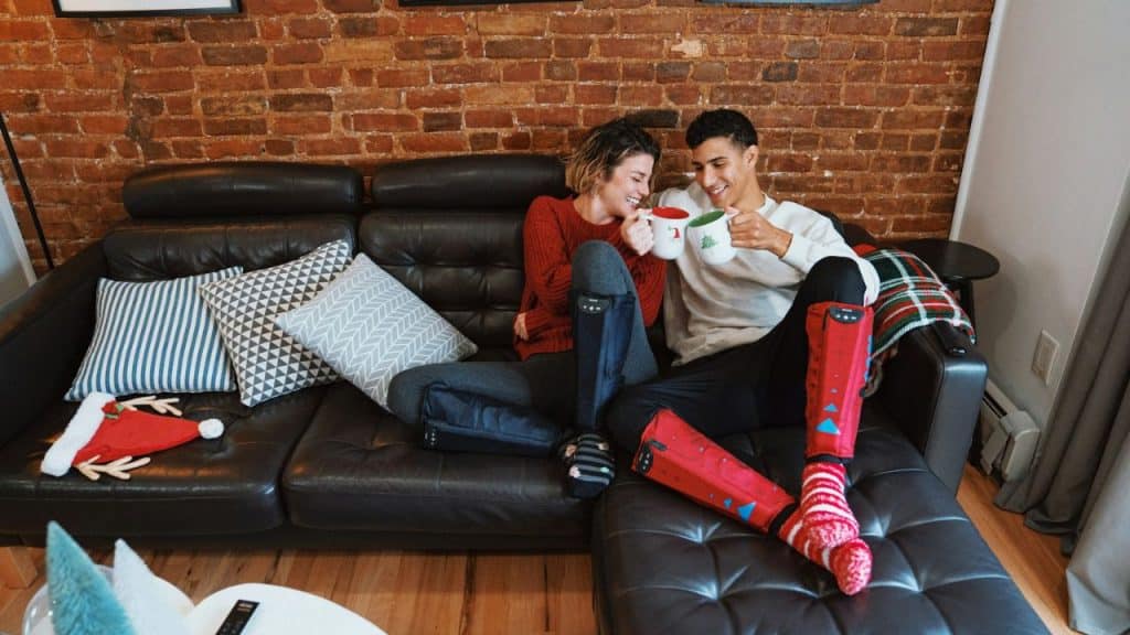 Couple on a black leather couch, wearing compression boots and toasting mugs.