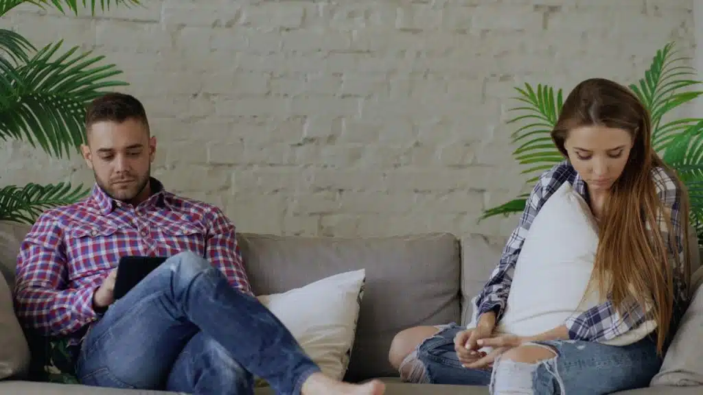 Couple sitting far apart on a couch; man uses a tablet, woman holds a pillow.