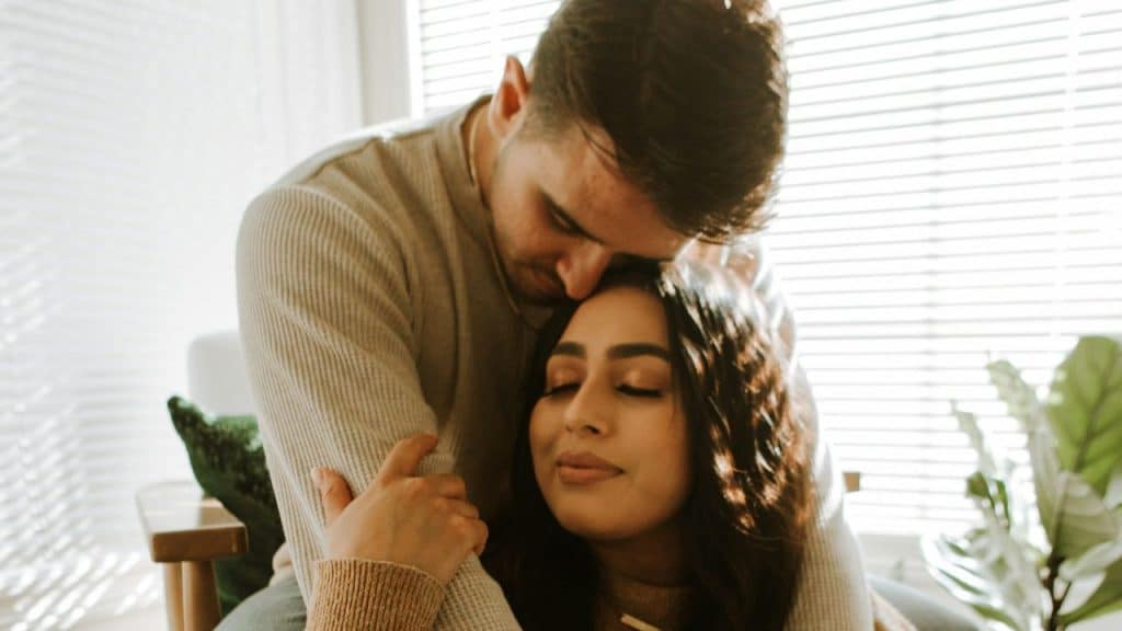 A young man embraces a woman from behind, resting his head on hers, near a window.