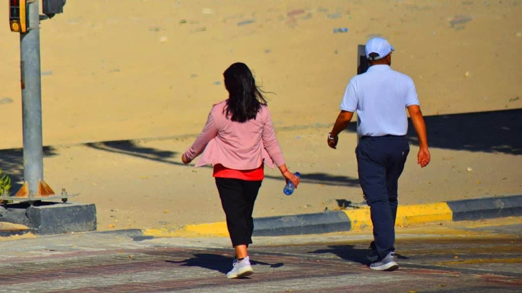 A couple walks away from the camera on a sidewalk near a sandy area.