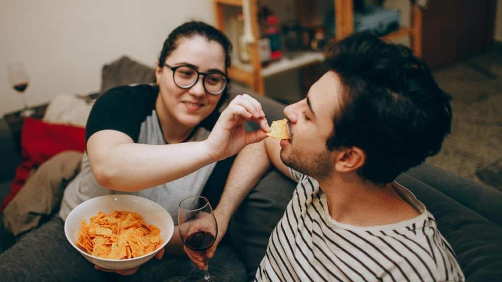A woman feeding a man a chip while they relax with snacks and wine.