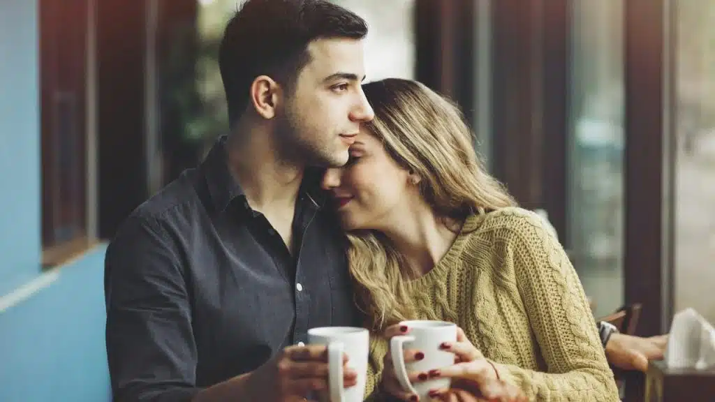 Woman resting her head on a man's shoulder while holding coffee cups indoors.