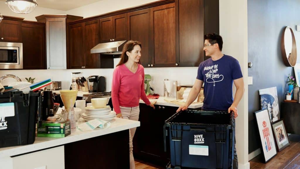 Man holding a moving box and woman standing in a kitchen with stacked dishes.