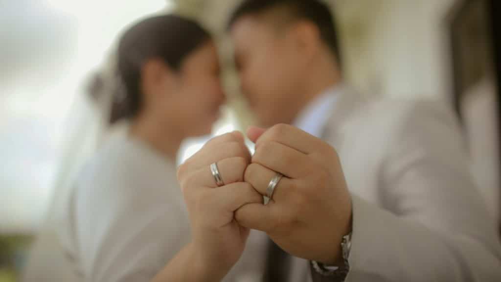 Close-up of a bride and groom touching fists, showcasing their wedding bands, with their faces out of focus in the background.