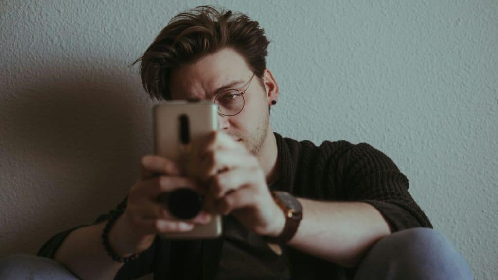 Man with glasses and dark shirt holding a phone, looking directly at the viewer.