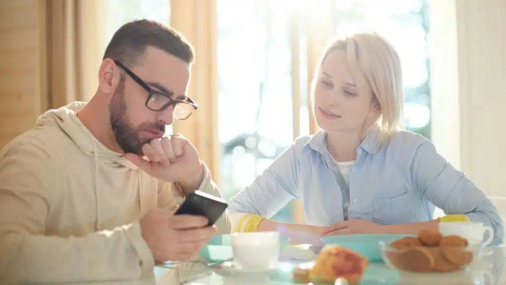 A man looks at his phone while a woman looks at him during a meal.