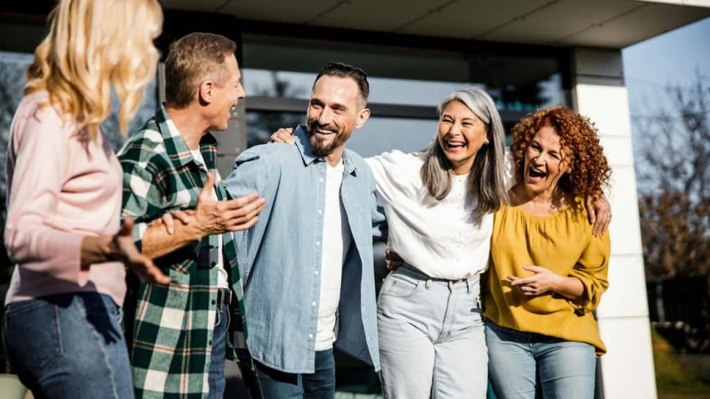 A diverse group of five smiling adults is standing closely together and laughing outdoors.