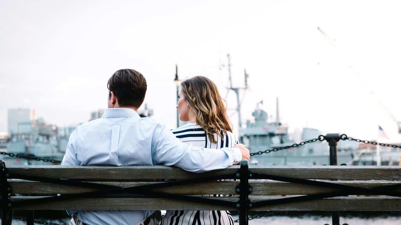 A couple sitting on a bench by the water with ships in the background.