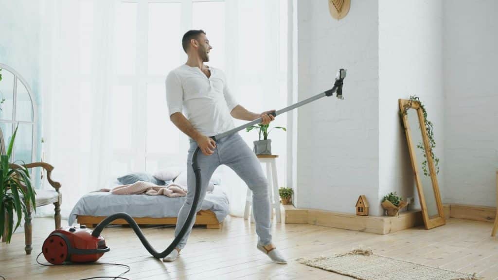 A man pretends a vacuum cleaner wand is a guitar while playfully dancing in a sunny room.