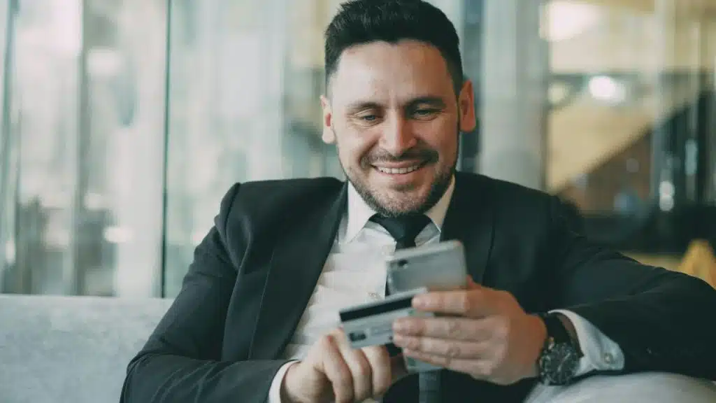 Man in a suit is smiling, holding a credit card and smartphone.