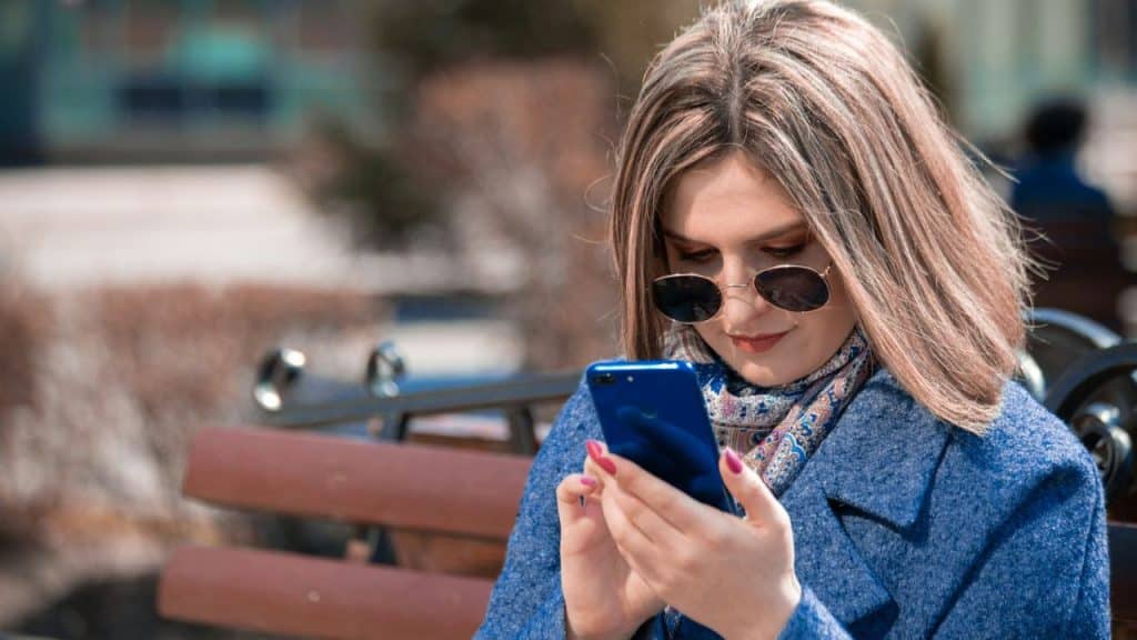 Woman with shoulder-length hair and sunglasses sitting outdoors, looking at her smartphone.