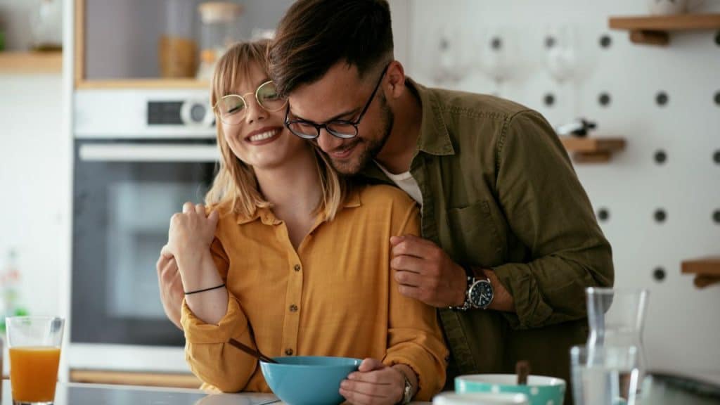 Smiling couple embracing in a kitchen, the woman eating cereal from a blue bowl.