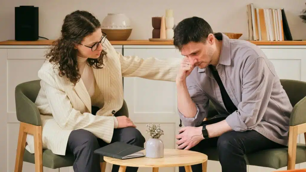A woman comforts a distressed man sitting across a low coffee table.