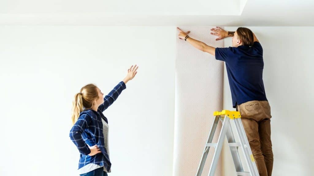 Man on ladder and woman applying patterned wallpaper to a white wall.
