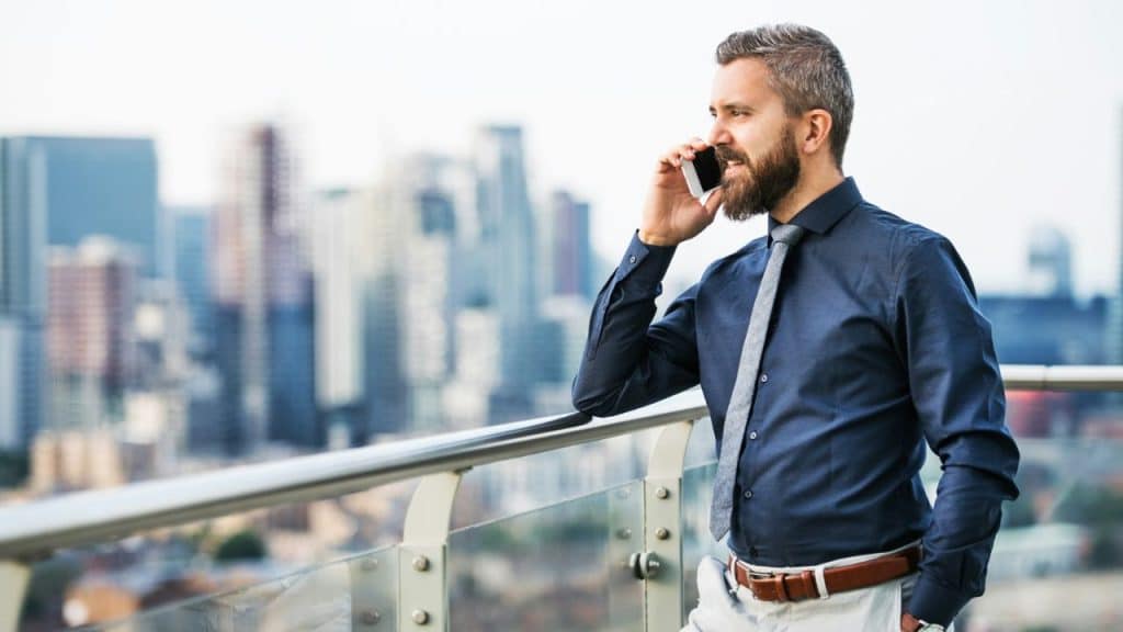 A bearded man in a blue shirt and tie talks on a cell phone on a city balcony.