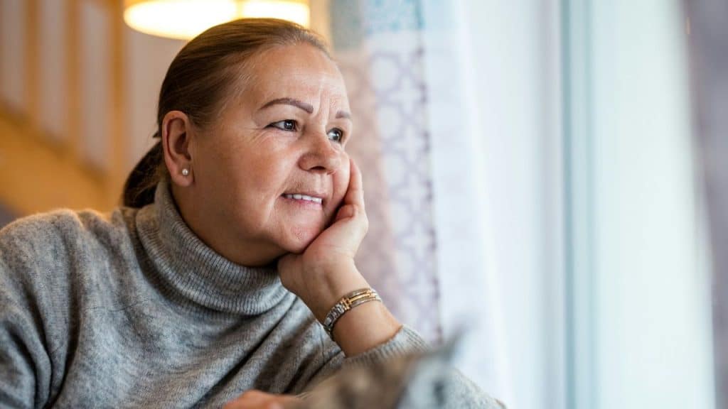 An older woman looks out a window, resting her chin on her hand.