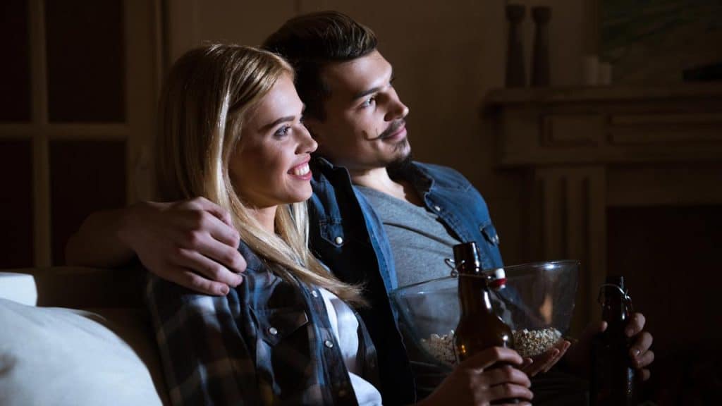 Couple watching TV with snacks and drinks.