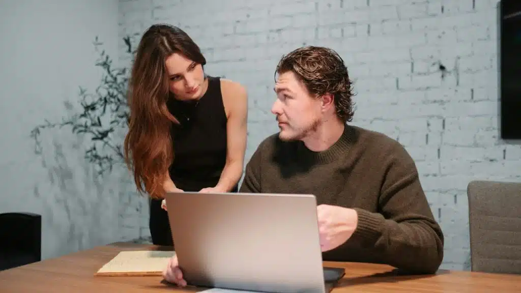 A man and a woman look at a laptop together at a wooden desk.