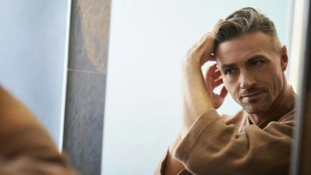 A man in a robe looks into a mirror, touching his styled hair.