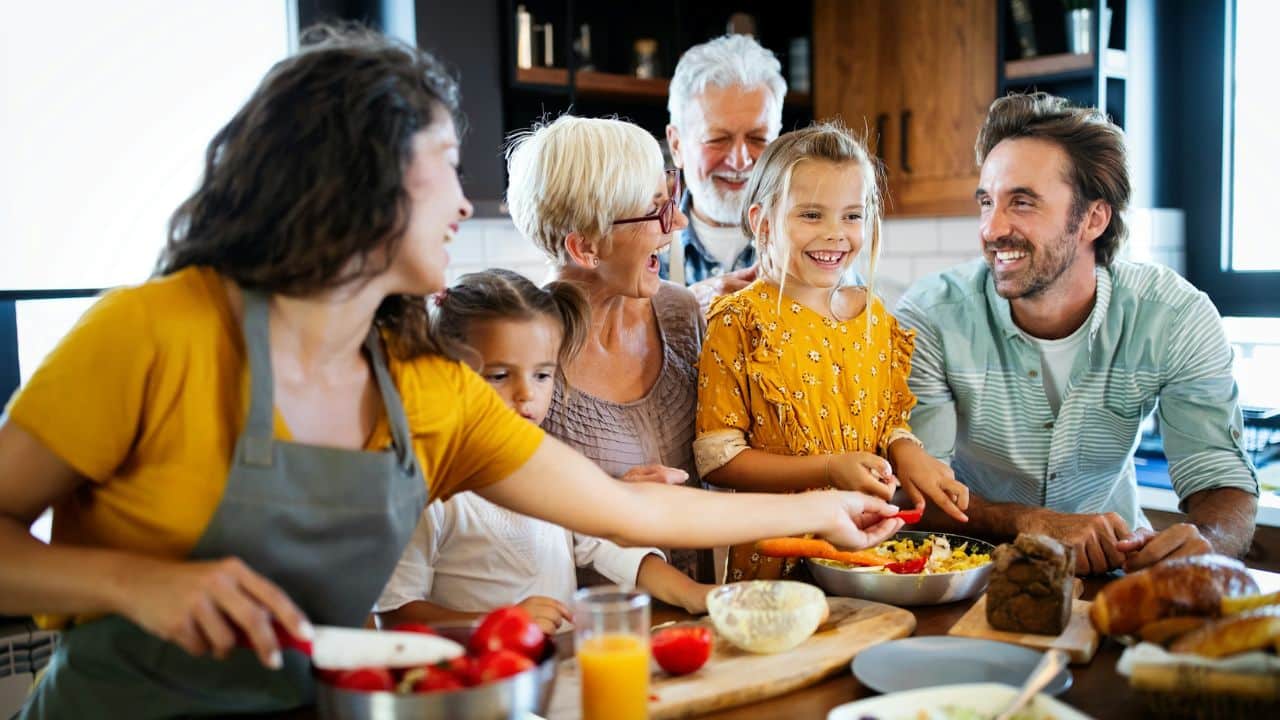 A multi-generational family, including grandparents and children, smiling around a kitchen table.