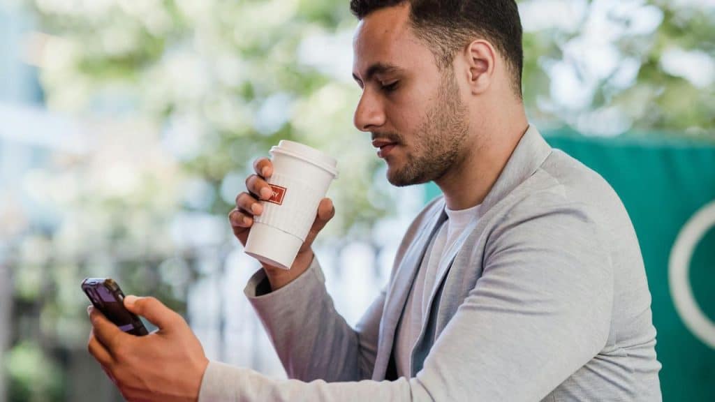 A man holding a coffee cup while looking at his phone outdoors.