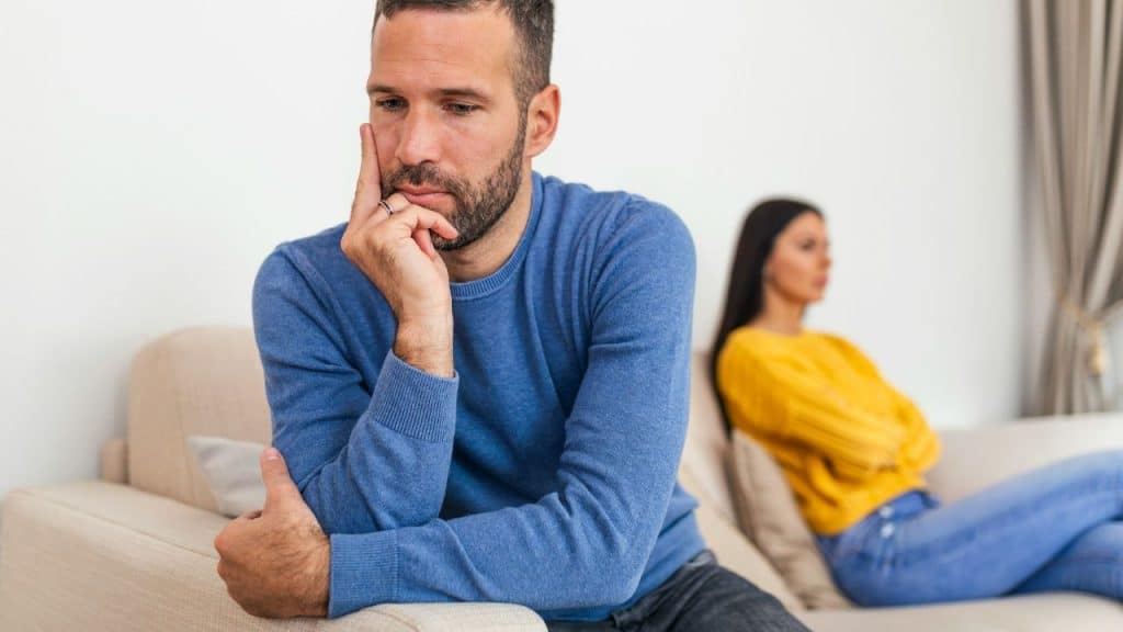 A man sits troubled on a couch in the foreground while a woman sits blurred and upset behind him.