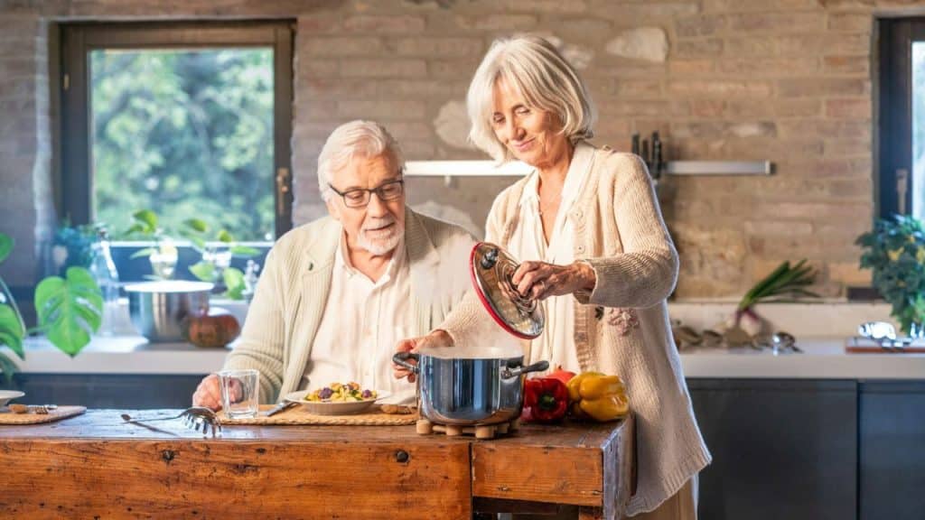 Elderly woman in a kitchen serving food to an elderly man sitting at a wooden table.