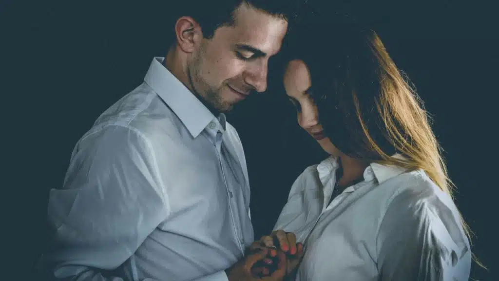 Young couple in white shirts stands close, holding hands, looking down.