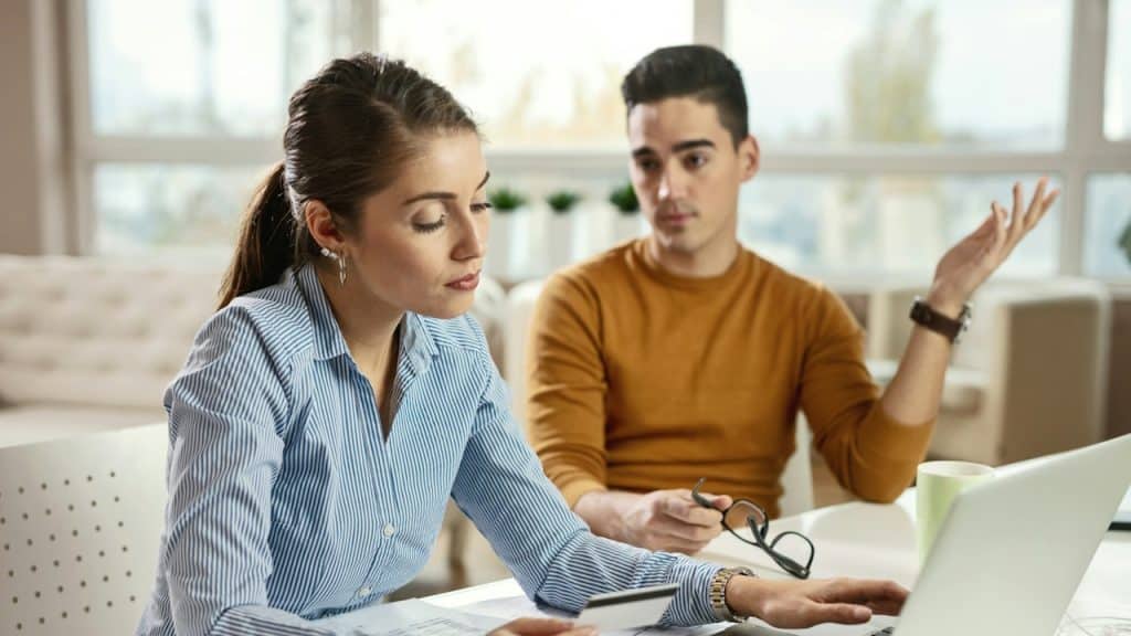 A man holding glasses and gesturing towards a woman who’s looking at her laptop.