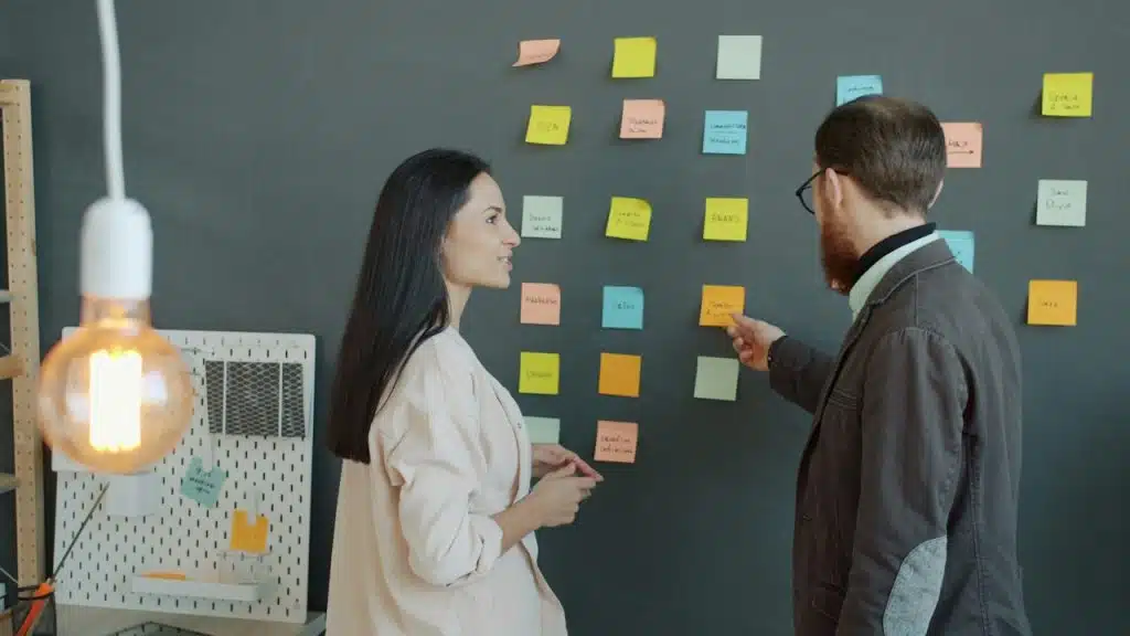 Two colleagues, a man and a woman, standing by a dark wall with colorful sticky notes, discussing ideas.
