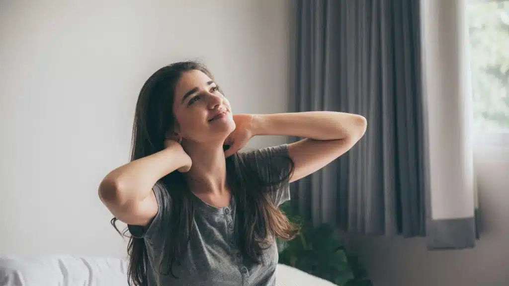 A woman sits on a bed smiling and stretching with her hands on her neck.