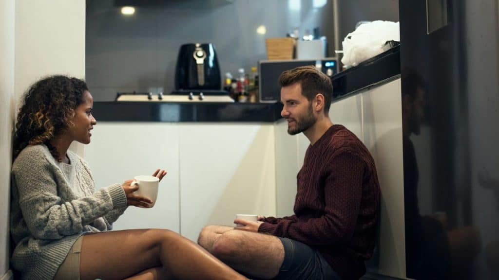A smiling couple sits on the floor in a kitchen, holding mugs and talking.