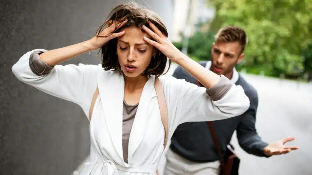 A woman clutches her head in distress as an arguing man approaches her on a sidewalk.
