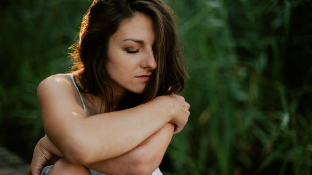 Woman sitting outdoors with eyes closed, hugging her knees against a green backdrop.