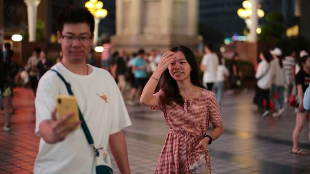 A man looks at his phone while a smiling woman walks beside him on a crowded street at night
